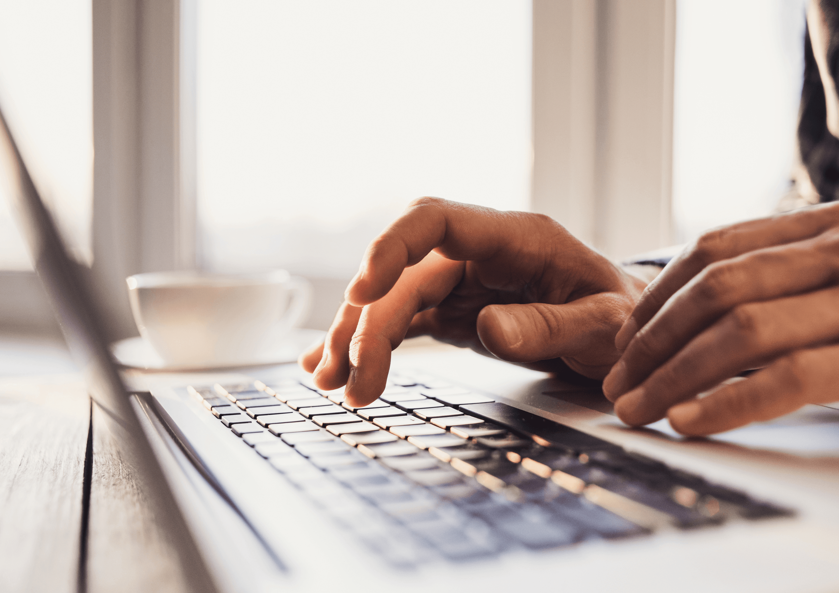 Close-up of hands typing on a laptop keyboard next to a cup of coffee